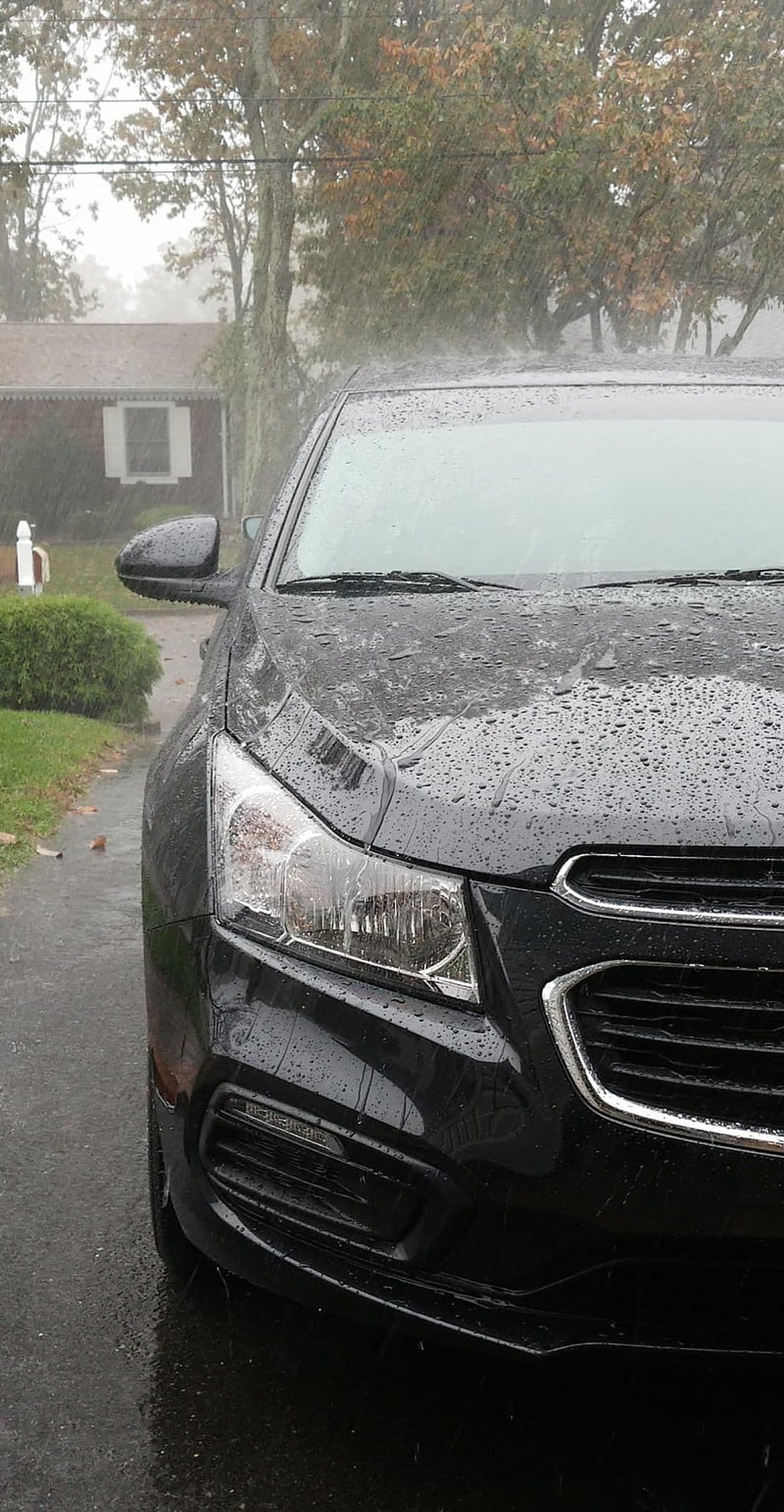 Black car hood covered in raindrops reflecting an overcast sky with visible headlights and front grille.