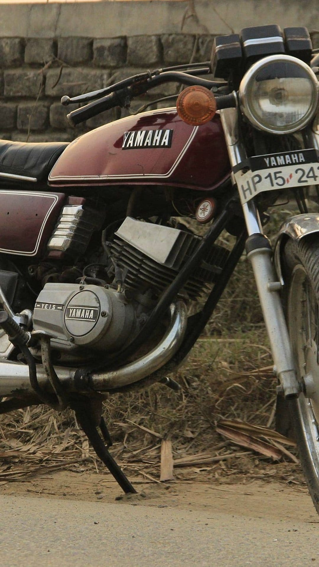 Maroon Yamaha RD350 motorcycle with chrome details parked on a dirt path next to a weathered brick wall.