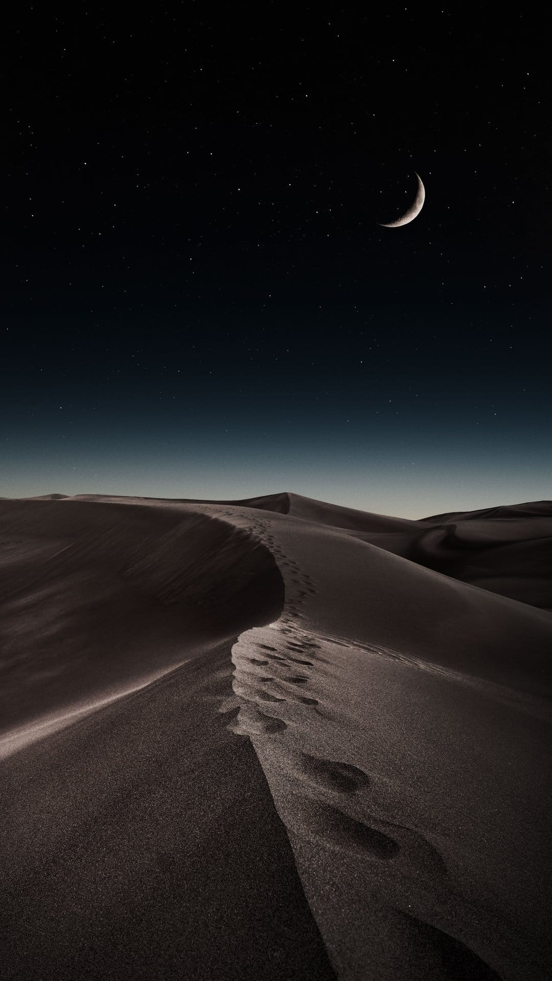 Footprints lead across dark sand dunes toward a crescent moon and a star-filled sky in a vertical night scene.