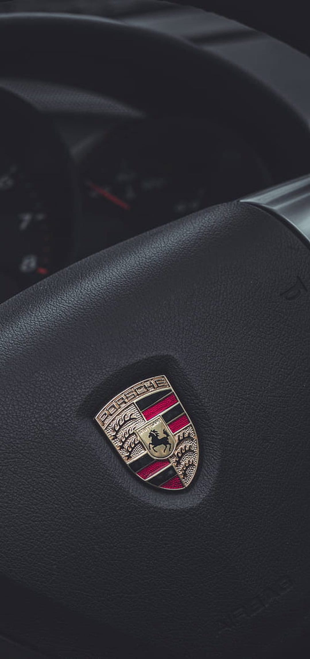 Porsche crest with gold shield and Stuttgart horse on a black leather steering wheel with visible stitching.
