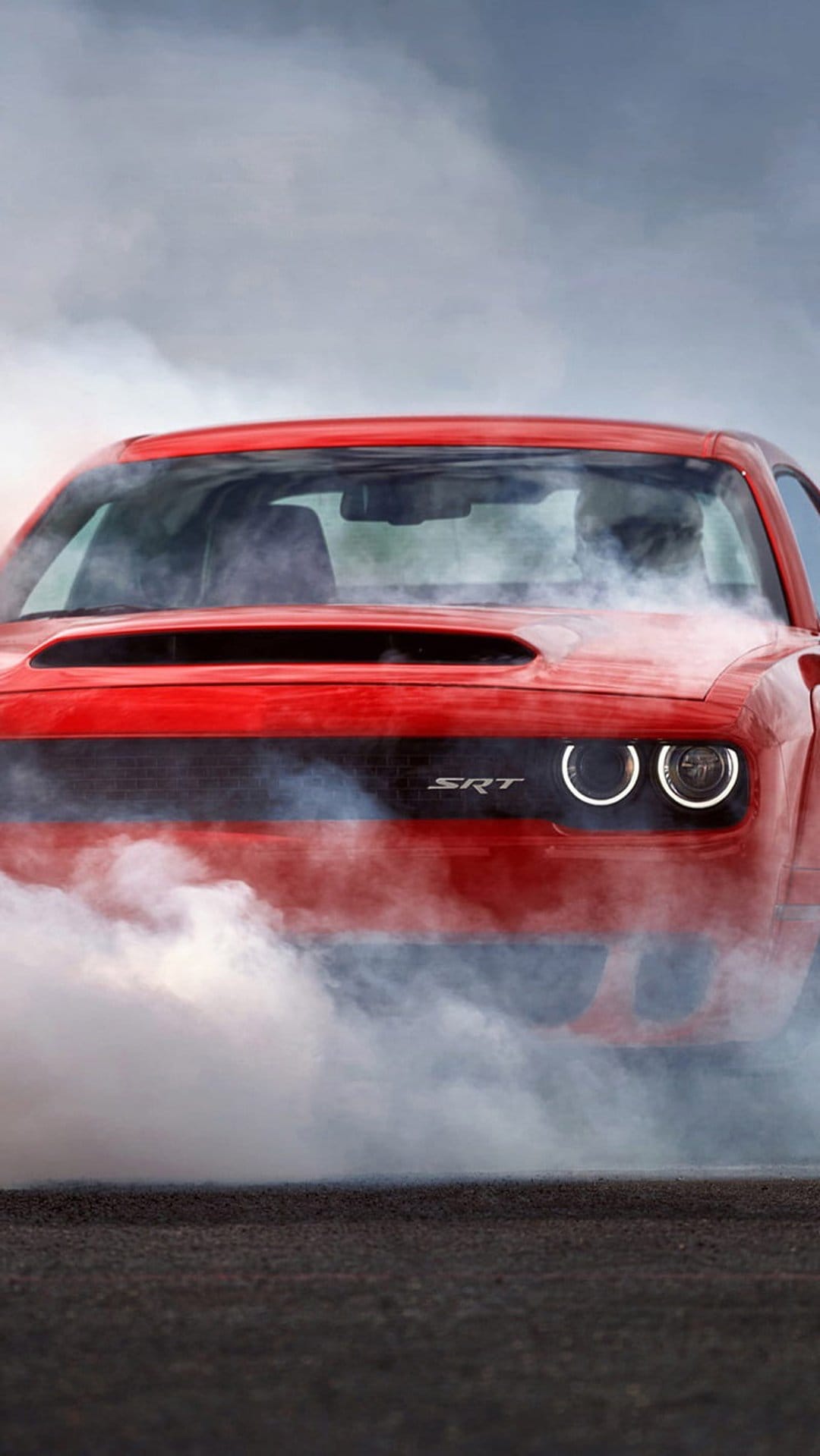 Red Dodge Hellcat muscle car emitting thick white tire smoke with glowing circular headlights and SRT grill logo.