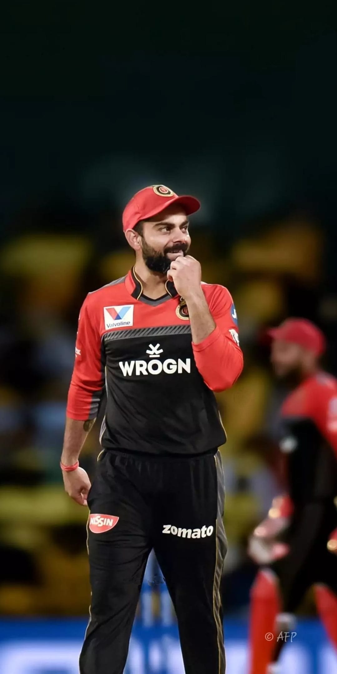 Virat Kohli smiling and touching his chin while wearing a red and black RCB cricket jersey and matching cap.