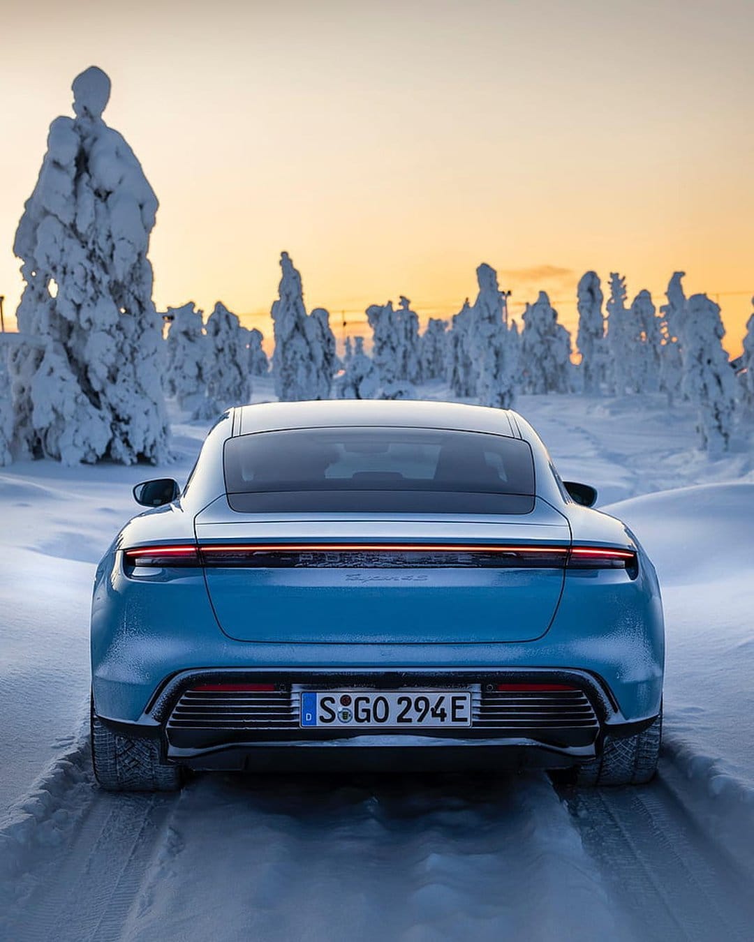 Blue Porsche Taycan rear view on a snowy road with pine trees and a warm orange sunset sky in the background.