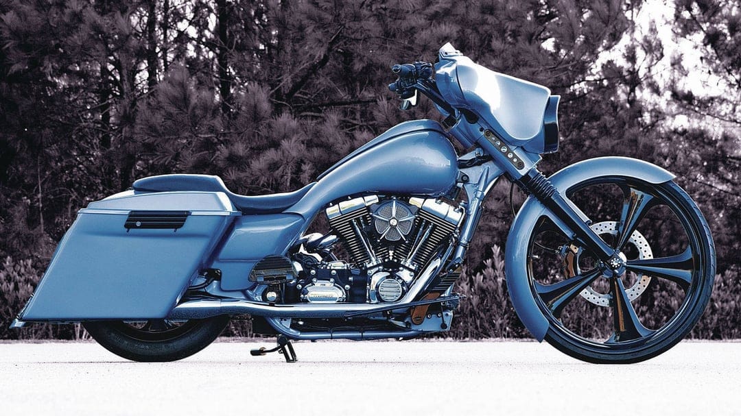 Blue custom bagger motorcycle with large black spoked wheels parked on a pavement before dark pine trees.