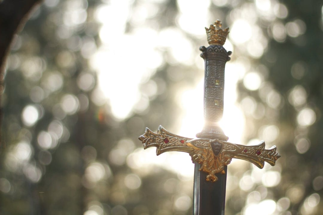 Crowned hilt of an ornate gold sword standing in a forest as sunlight filters through blurred autumn leaves.