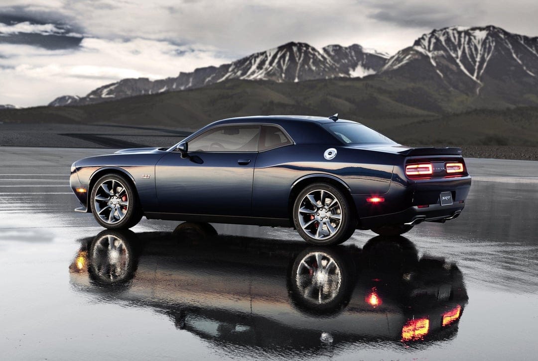 Dark blue Dodge Challenger SRT reflected on wet asphalt with snowy mountains in the background at dusk.