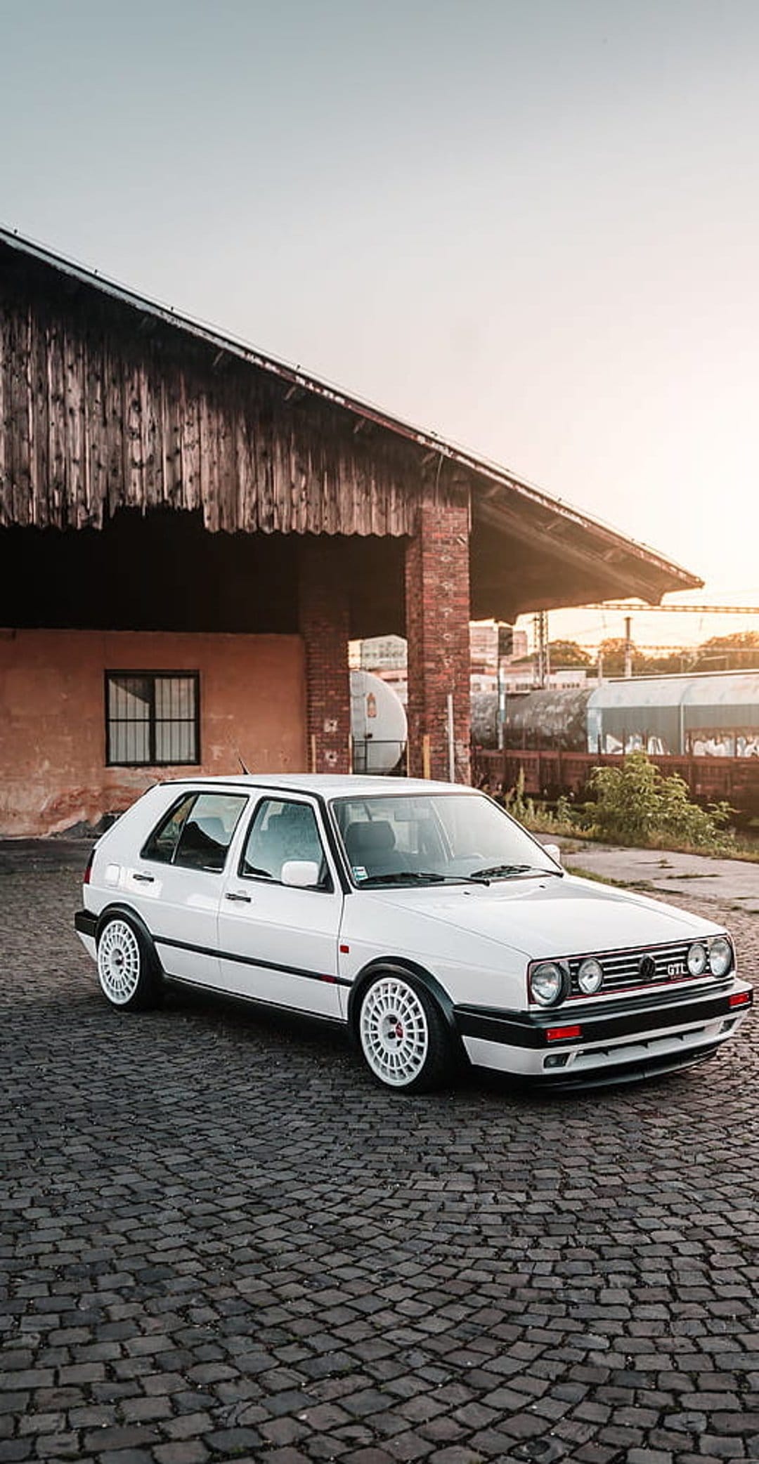 White Volkswagen Golf MK2 GTI parked on a cobblestone street in front of a rustic brick and wood building at sunset.