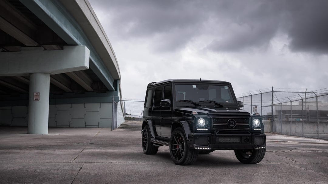 Black Brabus G-Class SUV with glowing LED lights parked on concrete under a bridge beneath a cloudy sky.