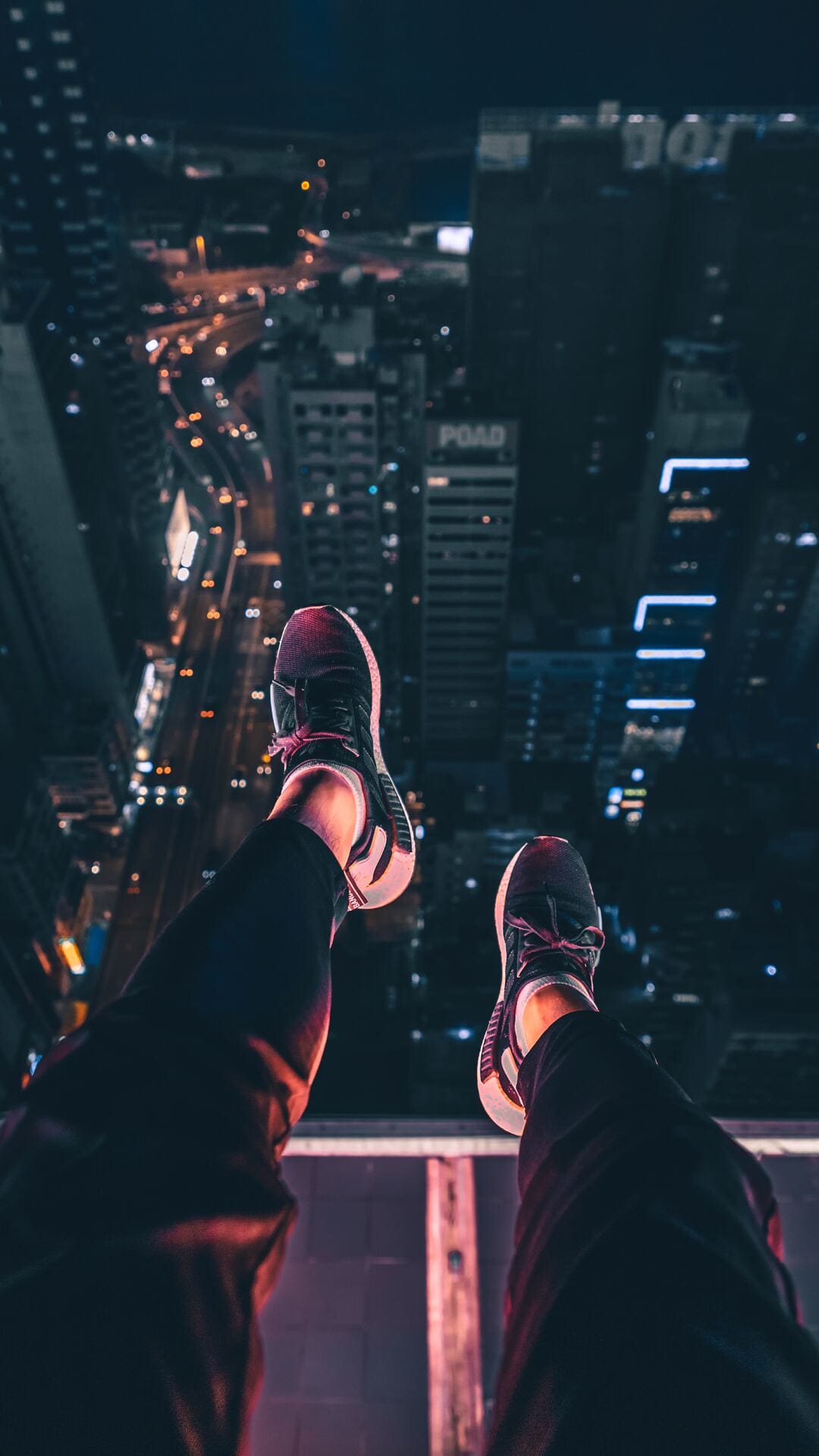 Sneakers dangle precariously over a skyscraper edge overlooking glowing city street lights at night.