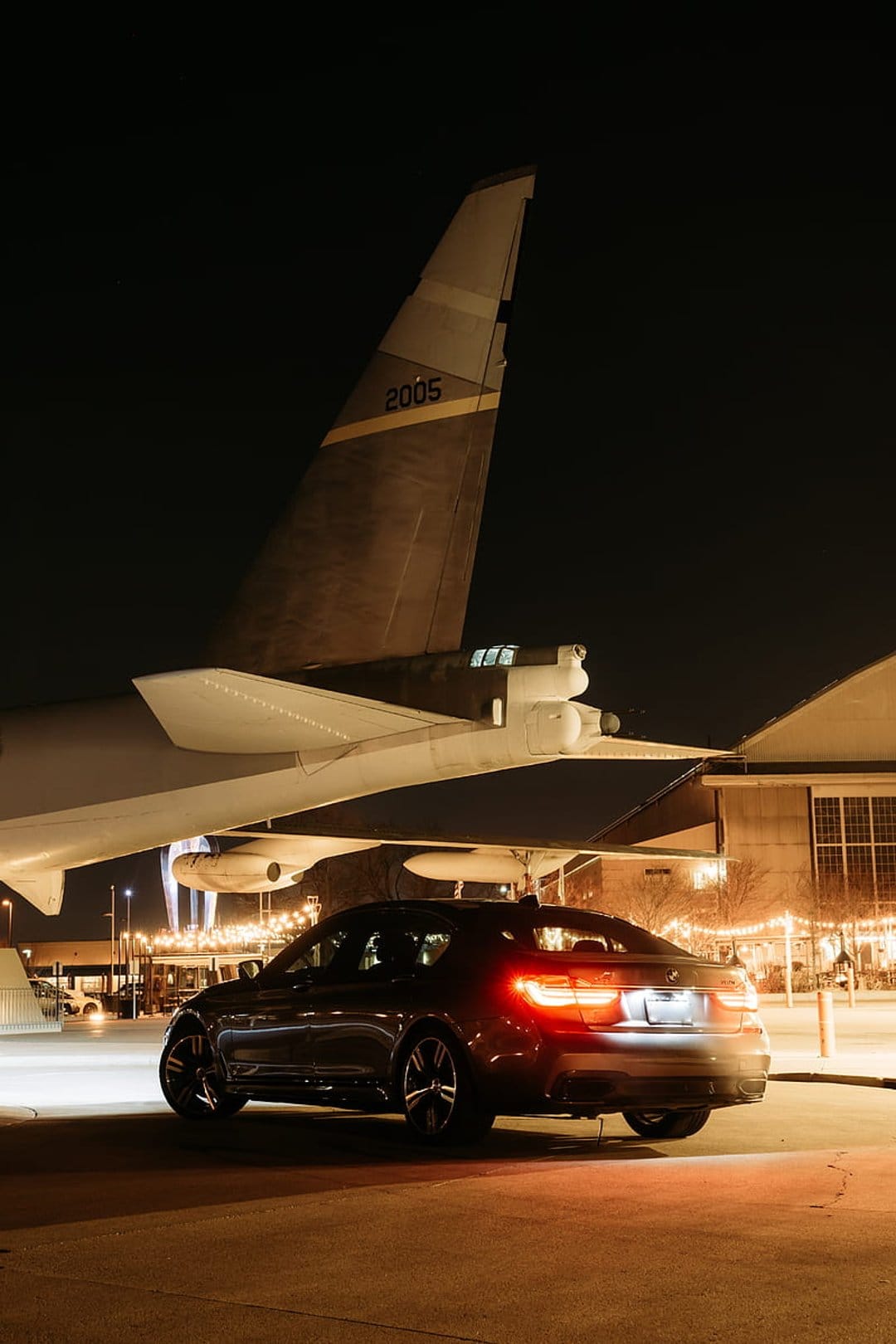 Luxury sedan with glowing red taillights parked on wet asphalt under the wing of a vintage airplane at night.