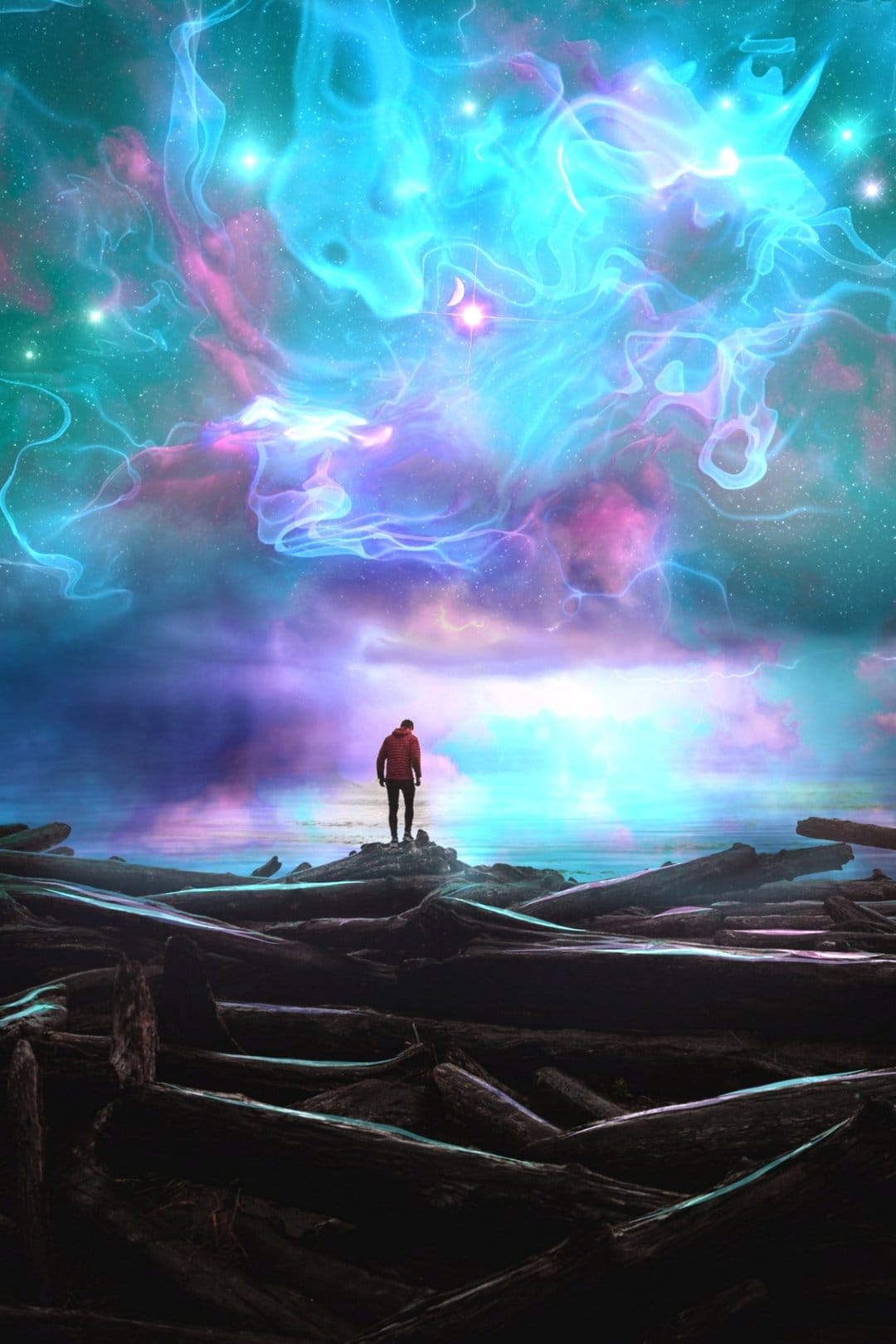 Man in a red jacket stands on driftwood watching a purple and blue cosmic nebula reflect in the ocean water.