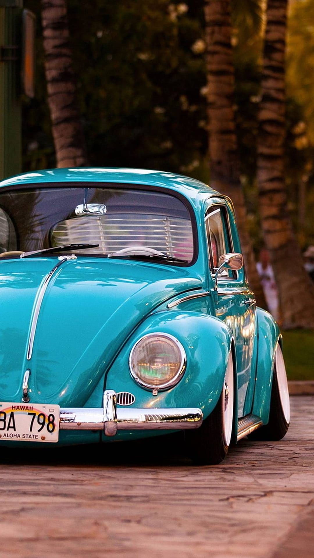 Turquoise Volkswagen Beetle with chrome accents parked on a wooden deck with blurred palm trees in the background.