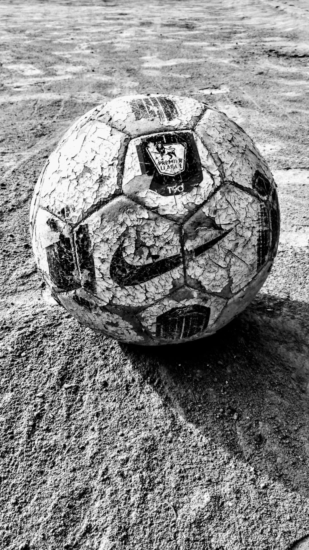 Worn Premier League soccer ball with peeling Nike logo resting on dusty, cracked ground in black and white.