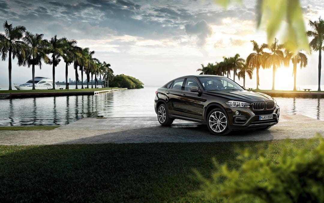 Luxury dark brown BMW X6 parked on a pier with palm trees and a yacht under a warm golden sunset sky.