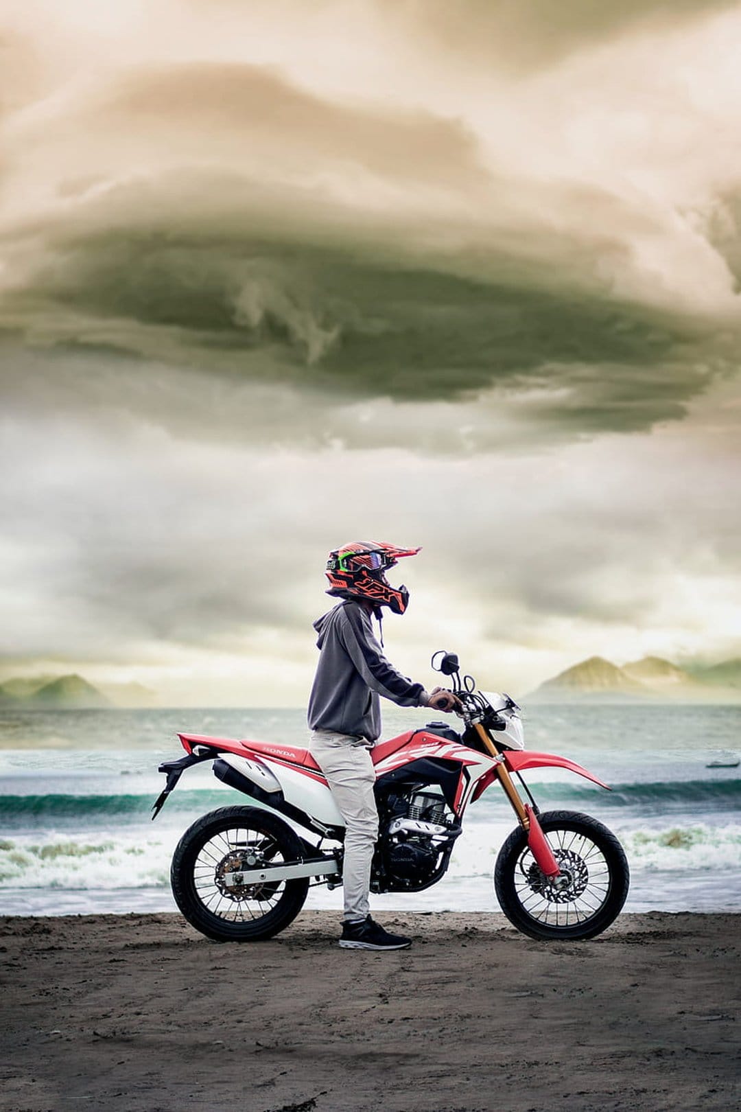 Motorcyclist in a red and white helmet stands by a dirt bike on a sandy beach under dark, stormy clouds.
