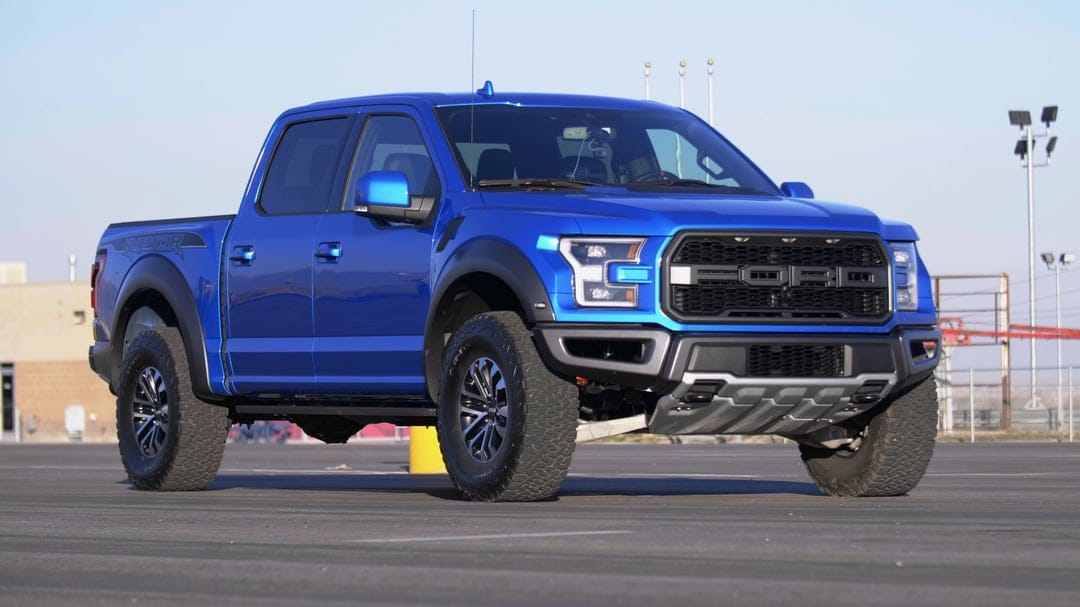 Blue Ford F-150 Raptor truck with off-road tires parked on an asphalt lot under a clear sky.