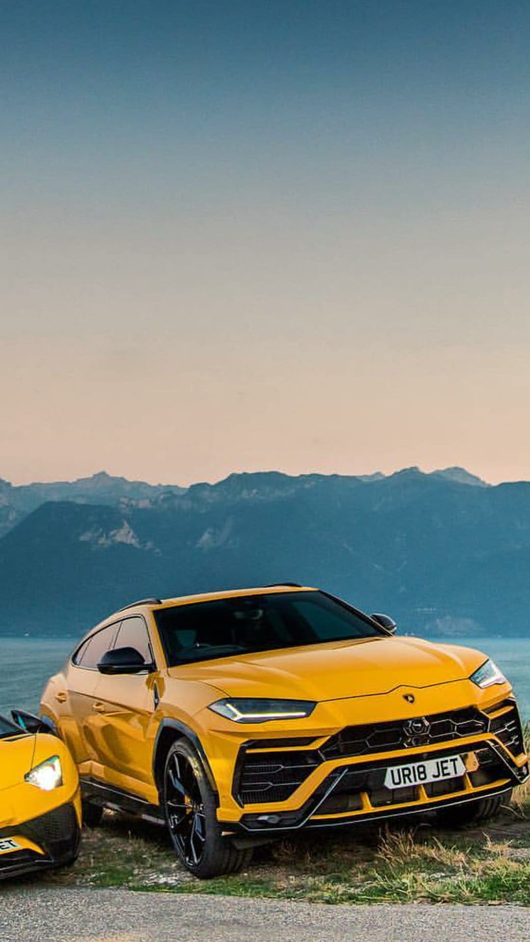 Two yellow Lamborghini Urus SUVs parked on a rocky shore with distant mountains and a calm lake background.
