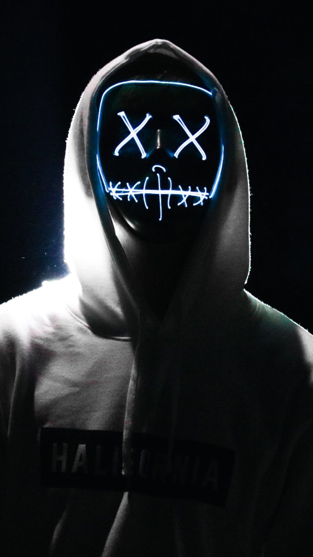 Hooded person wearing a glowing neon mask with X-shaped eyes and a sinister grin against a black background.