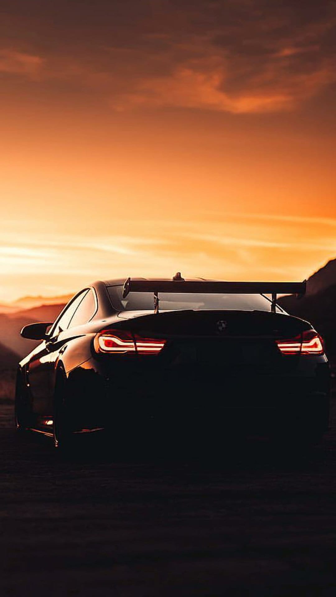 BMW M4 coupe silhouette with a large rear spoiler and glowing red taillights against a fiery orange sunset sky.