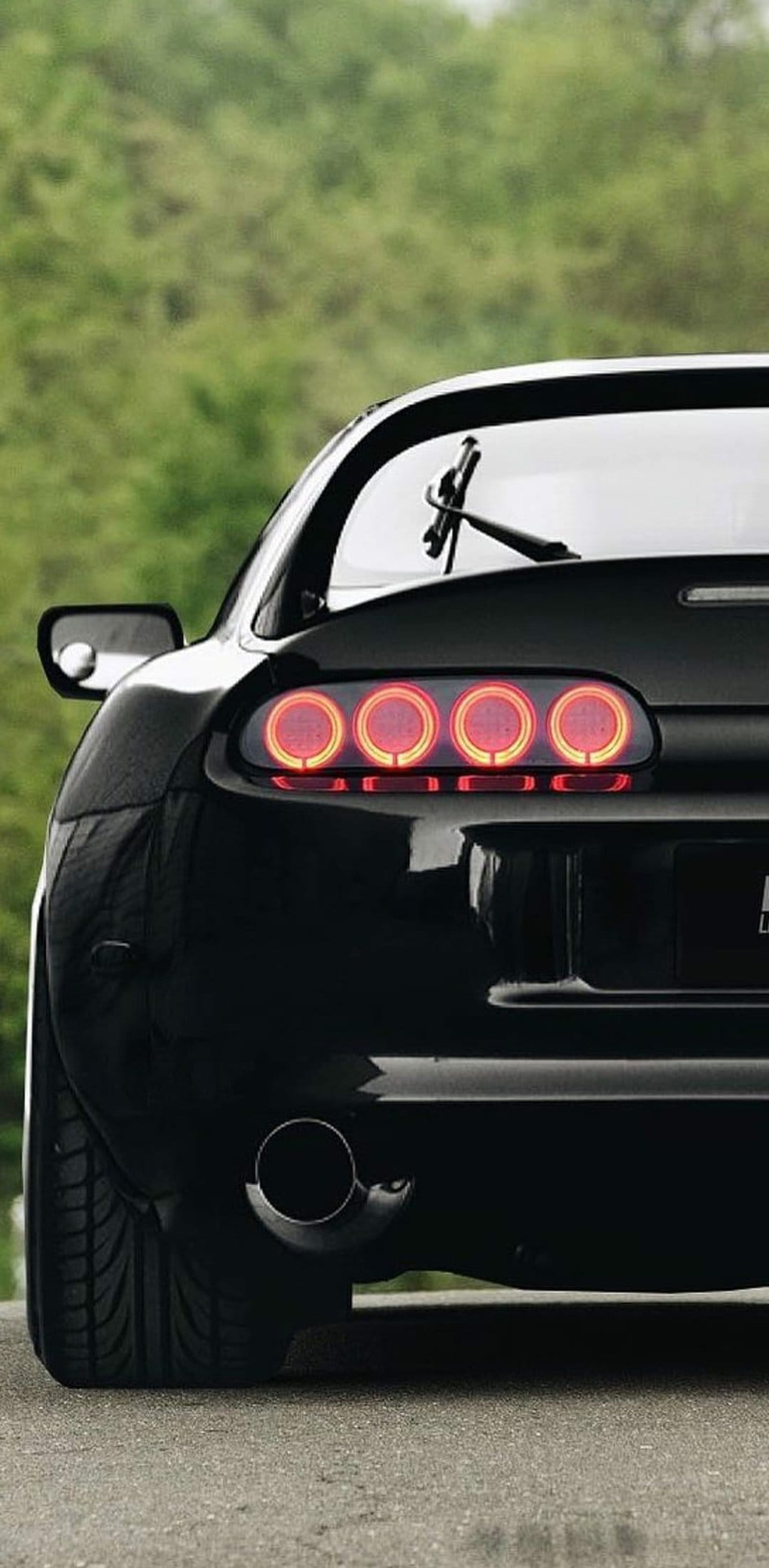 Black Toyota Supra rear view featuring four red circular taillights and a large chrome exhaust pipe outdoors.