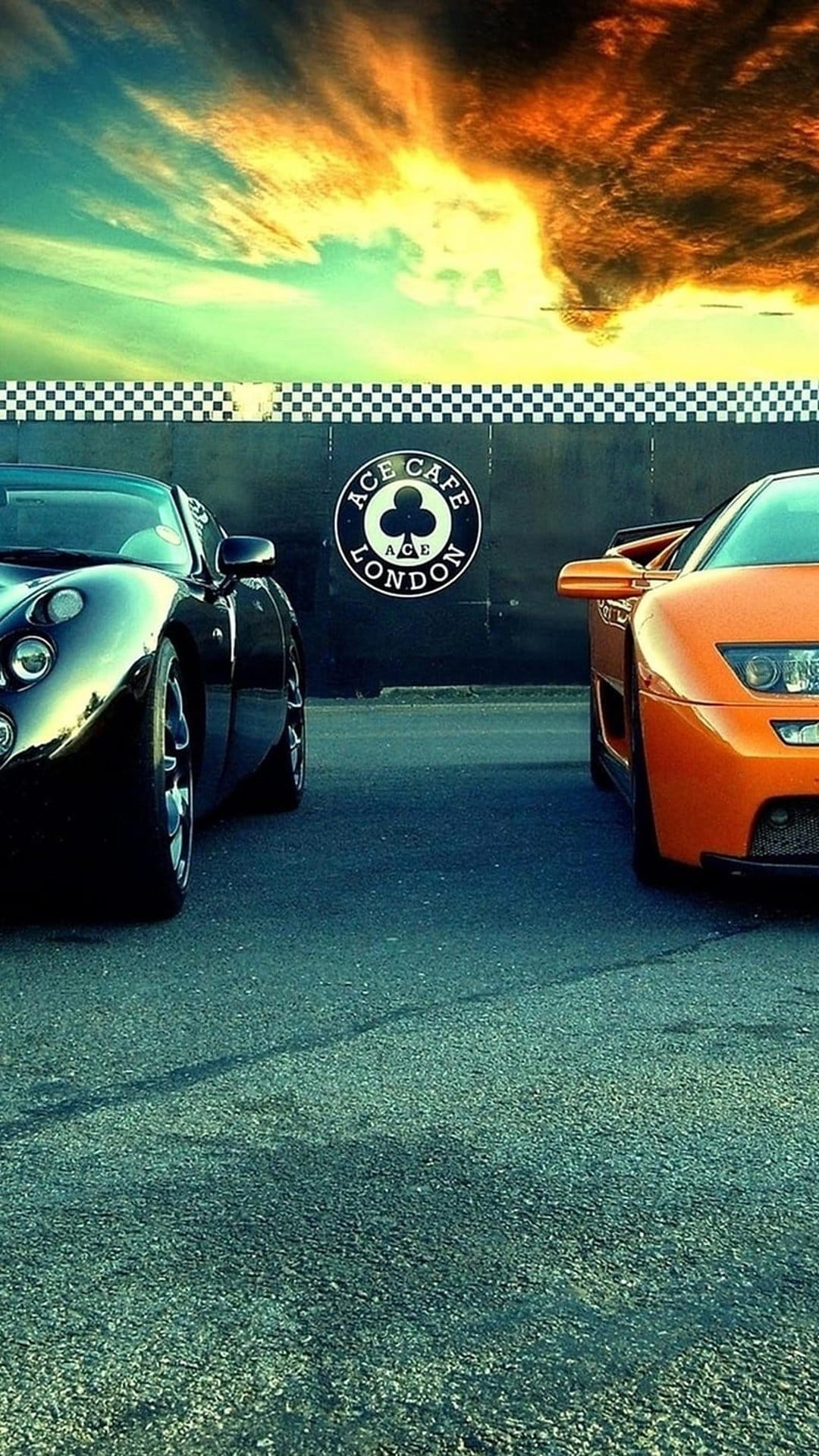 Black and orange sports cars parked in front of Ace Cafe London beneath a dramatic orange and turquoise sunset sky.