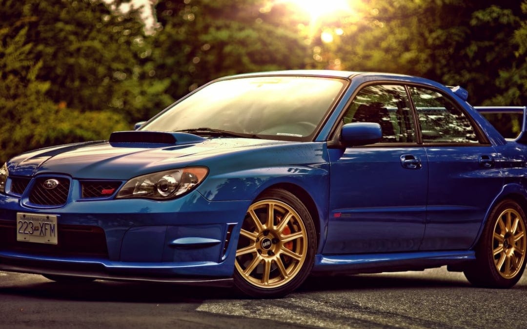 Blue Subaru WRX STI with gold wheels parked in a lush forest at sunset with warm light flares and bokeh background.