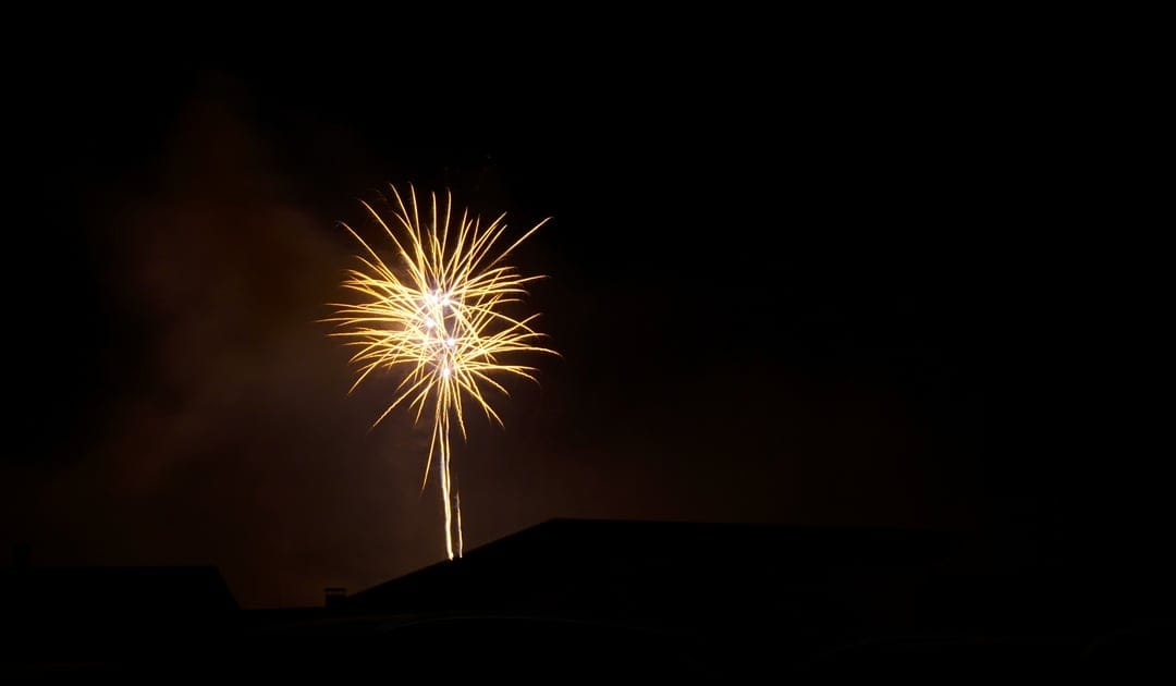 Golden starburst firework exploding in a dark night sky above black silhouettes of city rooftops and smoke.