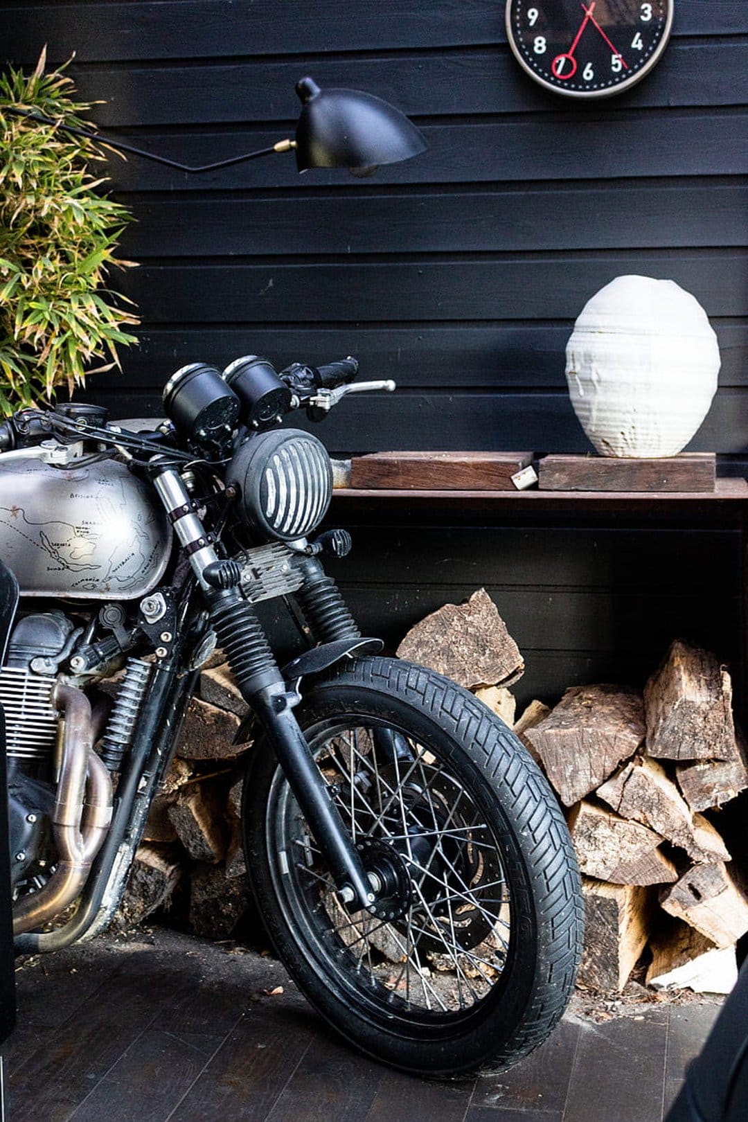 Vintage motorcycle parked next to a firewood pile and white vase against a dark wood wall with a wall clock.