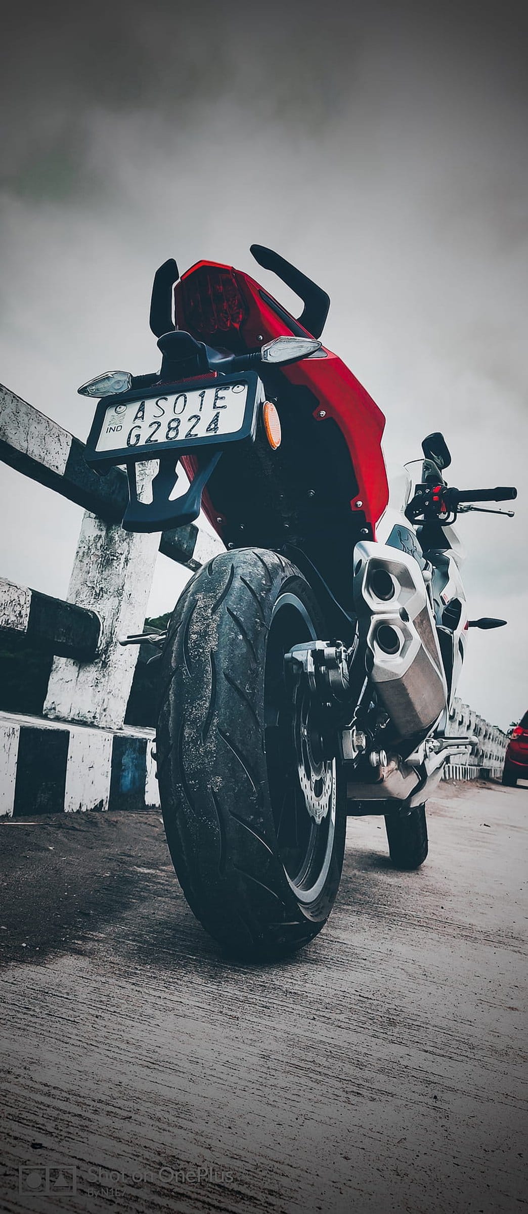 Red motorcycle rear view showing a wide tire, dual exhausts, and a lit license plate against a concrete barrier.