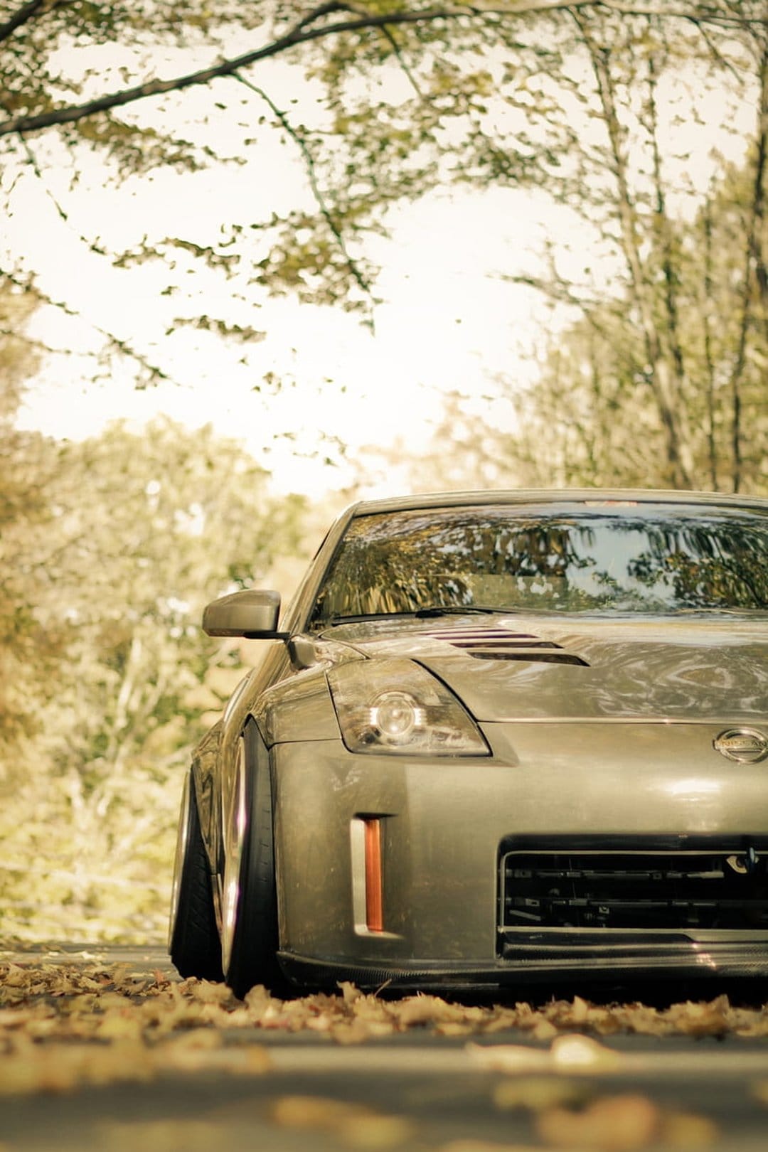Silver Nissan 350Z sports car on a road covered in orange autumn leaves with tree reflections in the windshield.