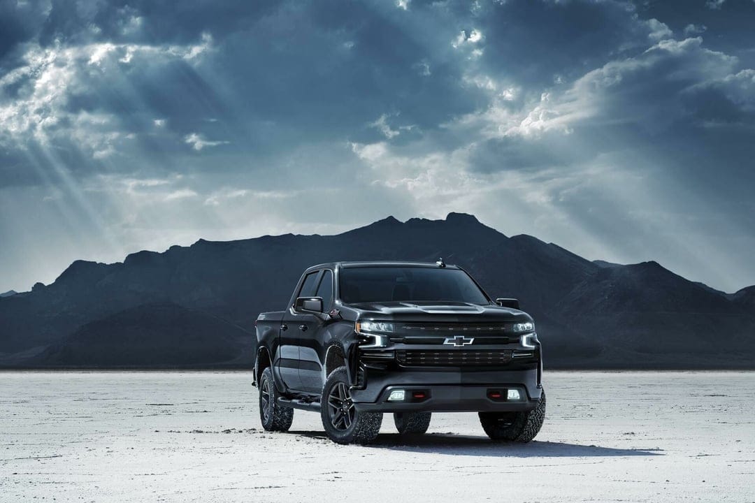 Black Chevrolet Silverado pickup truck on a white salt flat under a moody cloudy sky with dramatic sun rays.