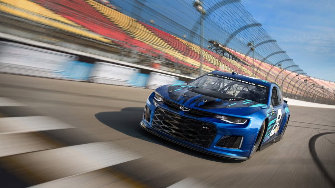 Blue Chevrolet Camaro ZL1 race car speeding on an oval track with motion-blurred grandstands and blue sky.