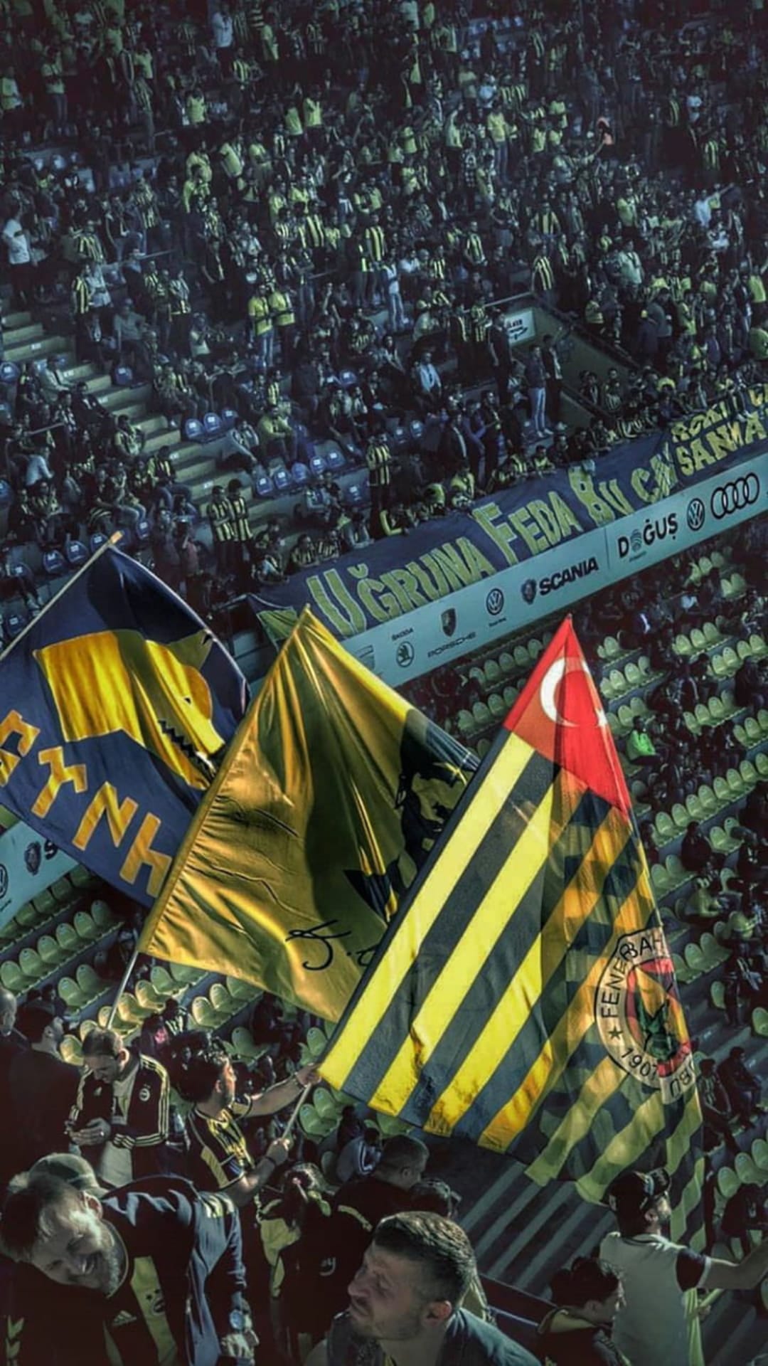 Fenerbahçe fans wave yellow and navy blue flags in a packed football stadium alongside a Turkish national flag.