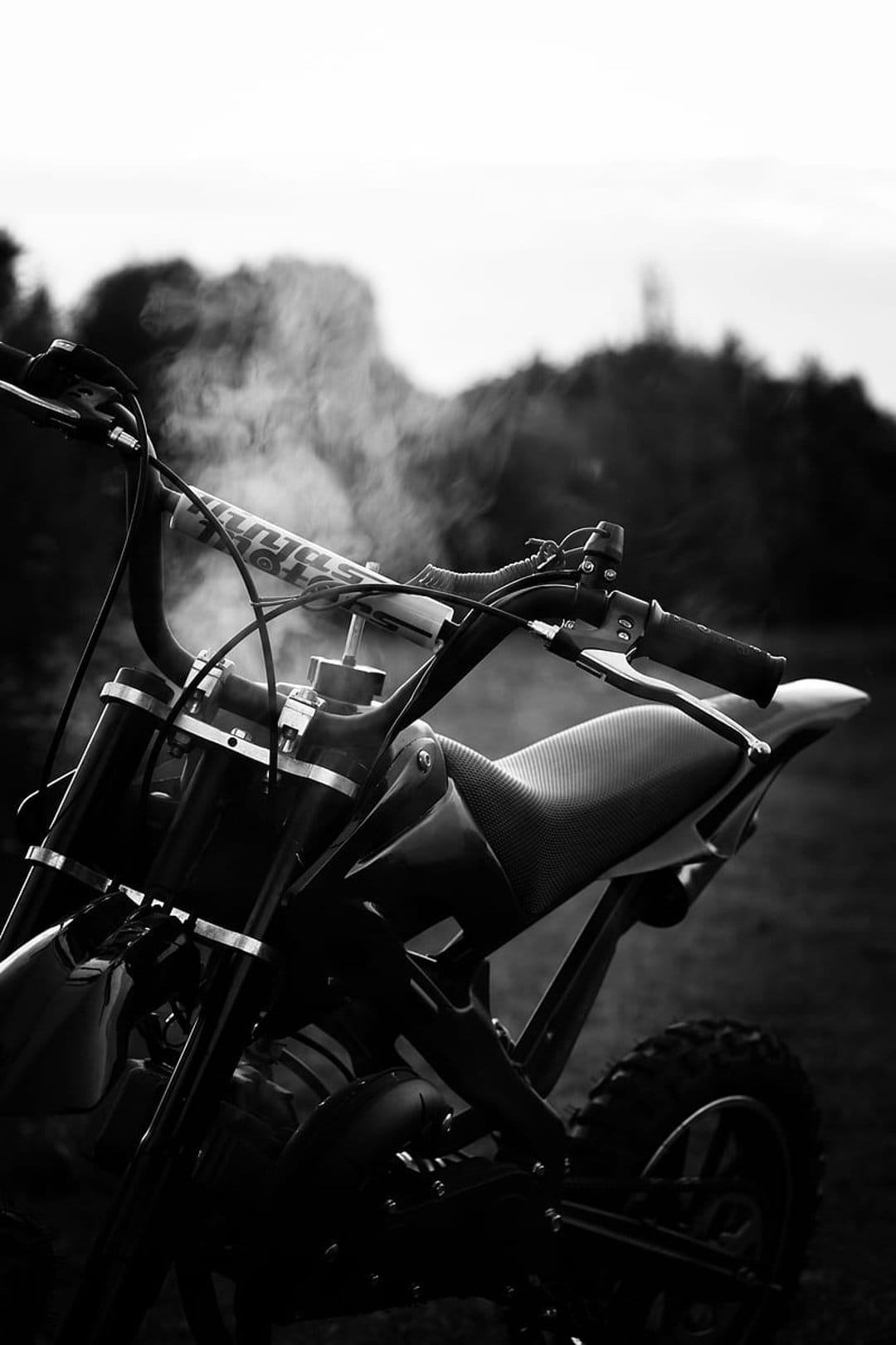 Dirt bike handlebars and a textured seat are framed by rising engine smoke in a monochrome outdoor setting.