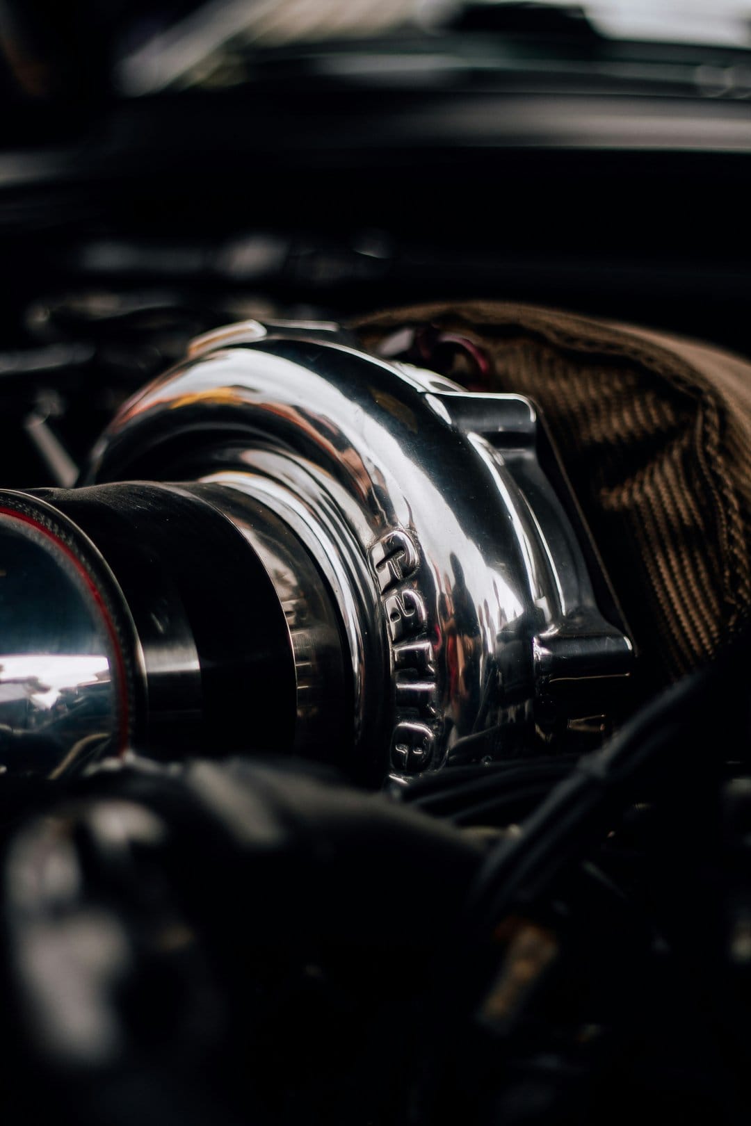 Chrome turbocharger housing with polished metal surfaces and intake piping against a blurred engine bay background.