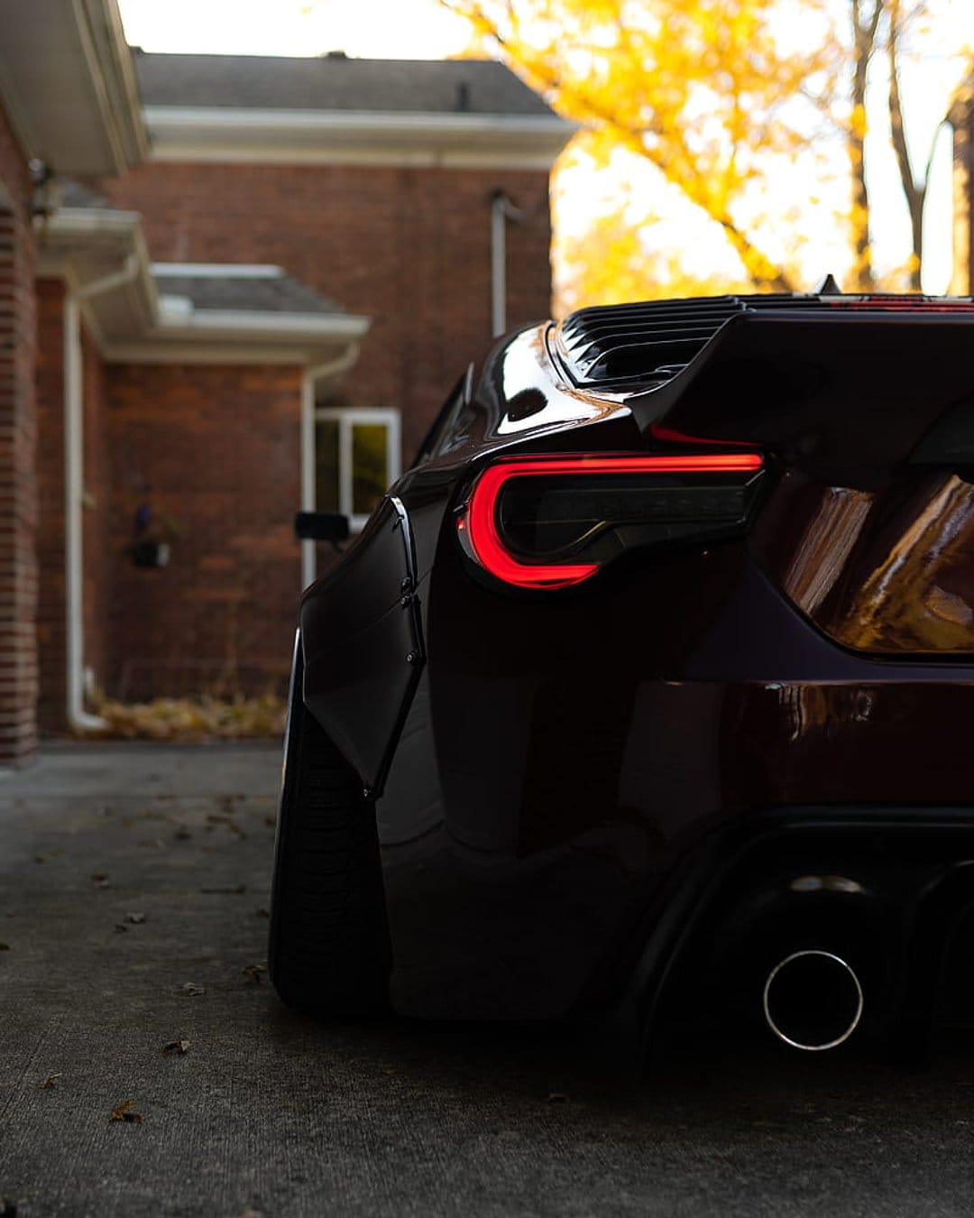Dark red sports car rear with glowing C-shaped LED taillights, a large spoiler, and autumn leaves behind it.