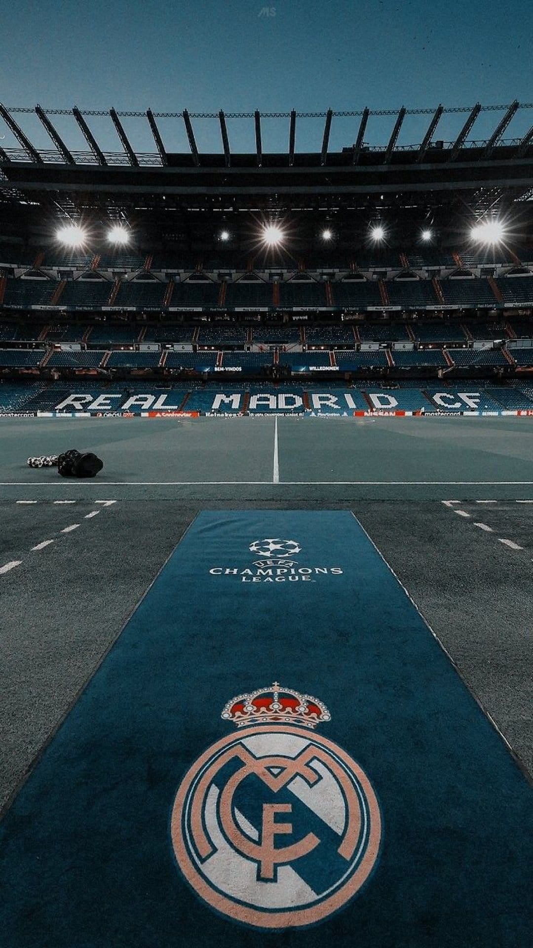 Real Madrid crest and Champions League logo on a blue carpet leading to an illuminated stadium at night.