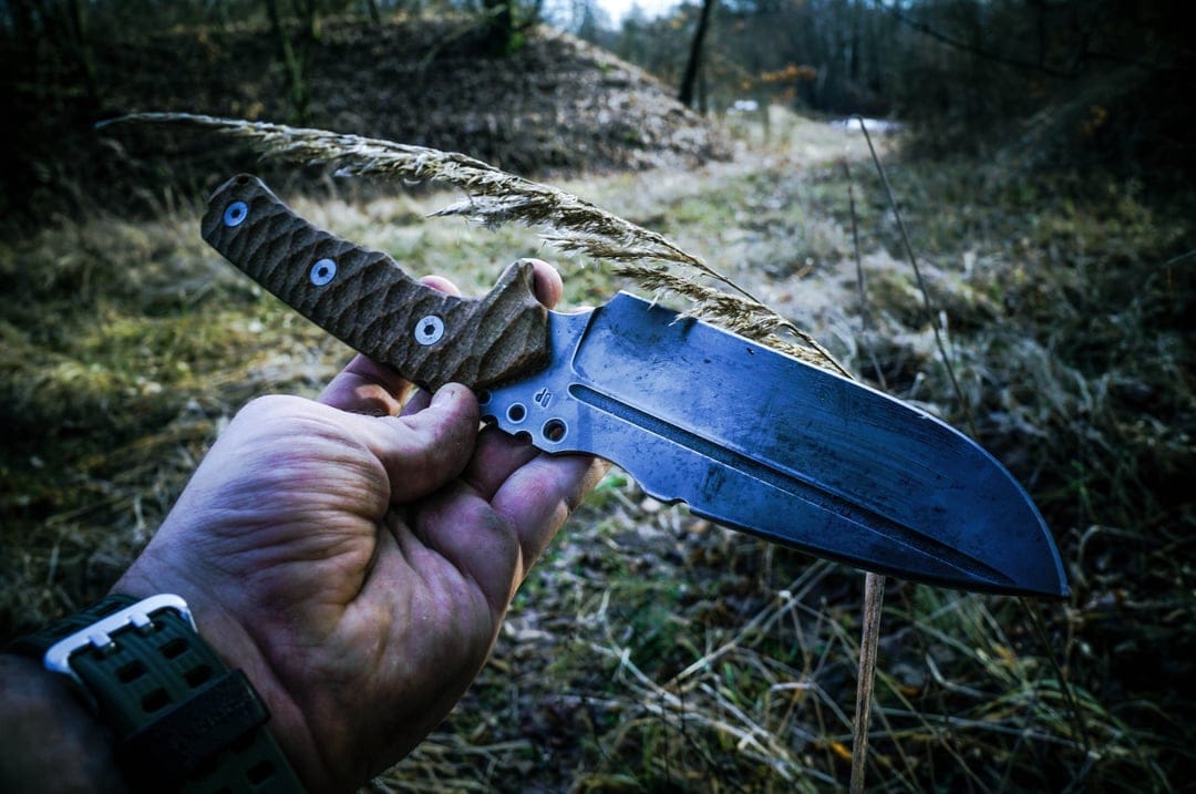 Man's hand grips a tactical steel knife with a textured handle against a blurred autumn forest background.
