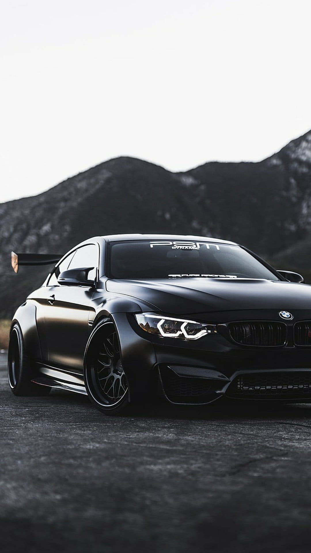 Matte black BMW M4 widebody coupe with a large rear spoiler and halo headlights on a blurred mountain road.