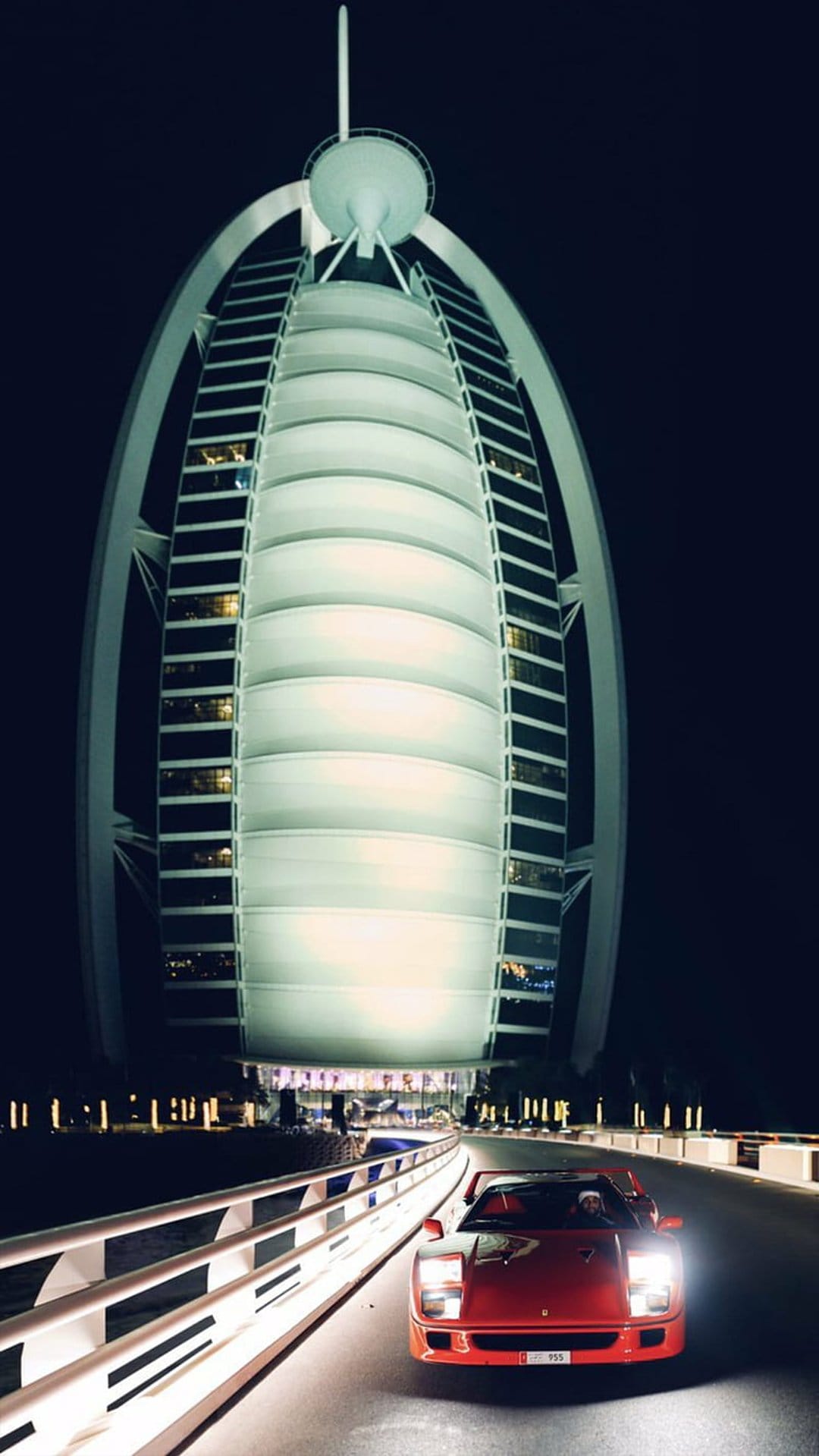 Red Ferrari sports car driving on a road at night with the sail-shaped Burj Al Arab hotel glowing in the background.