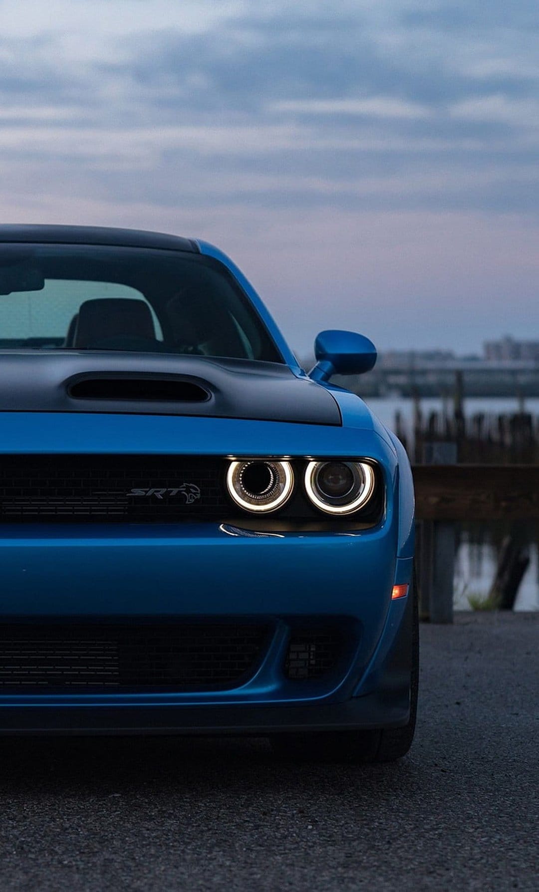 Dodge Challenger SRT Hellcat headlights glow with circular white LEDs on a blue car front at twilight near water.