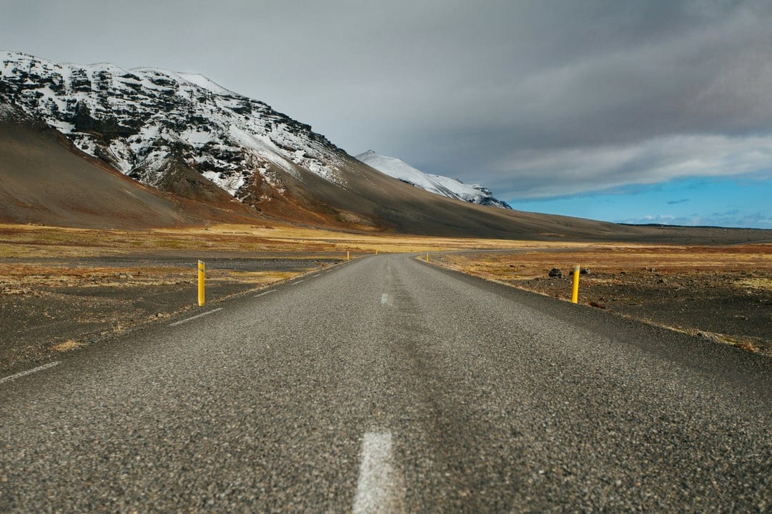 Long asphalt road with yellow markings leading toward snow-capped mountains under a dark cloudy sky.