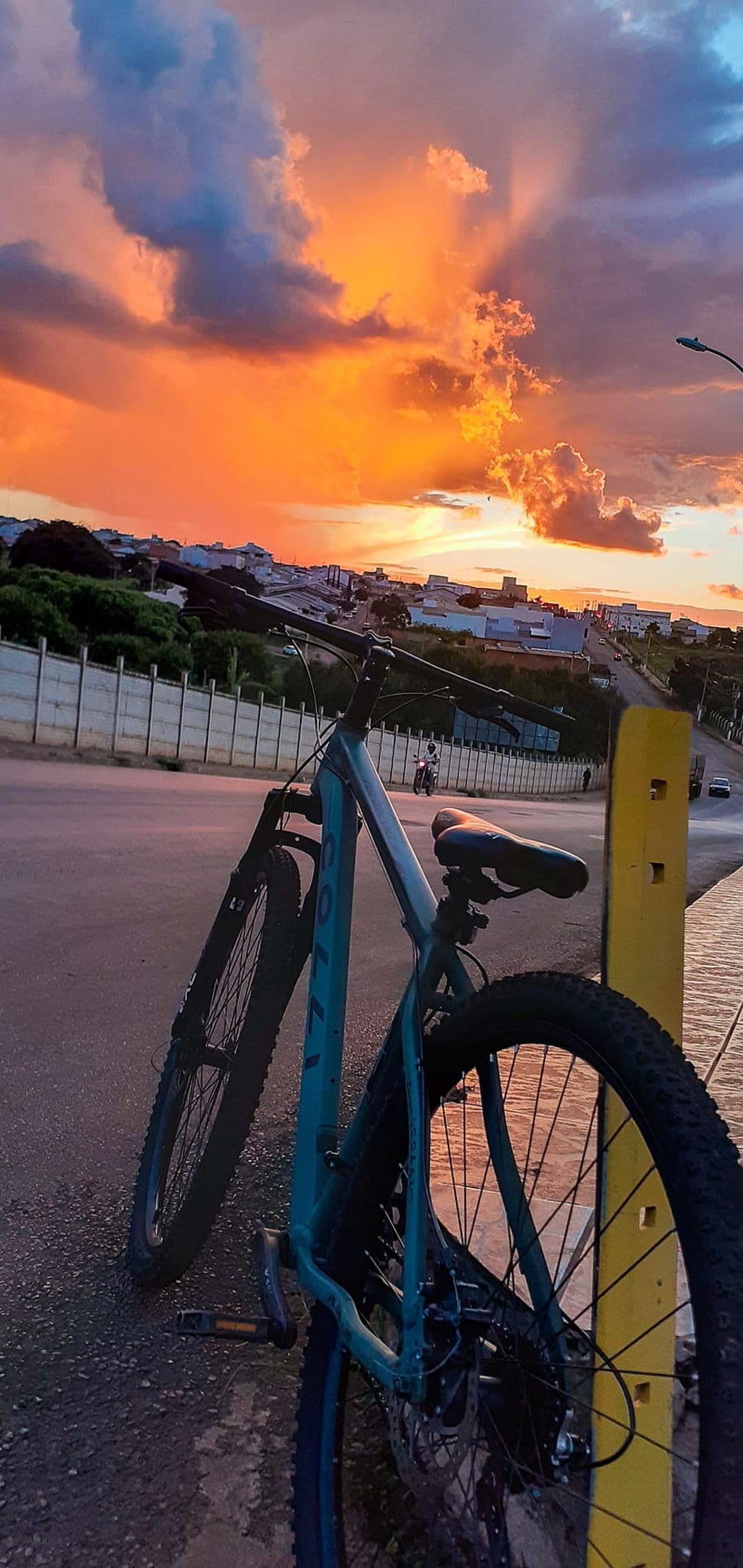Teal bicycle leaning on a yellow post during a purple and orange sunset over a distant city skyline at dusk.