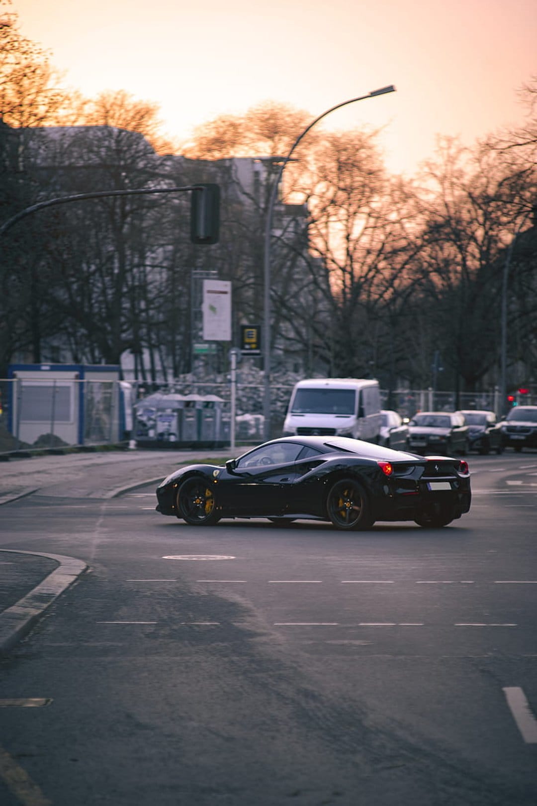Black Ferrari sports car turns onto a city street during a vibrant orange sunset with trees in the background.