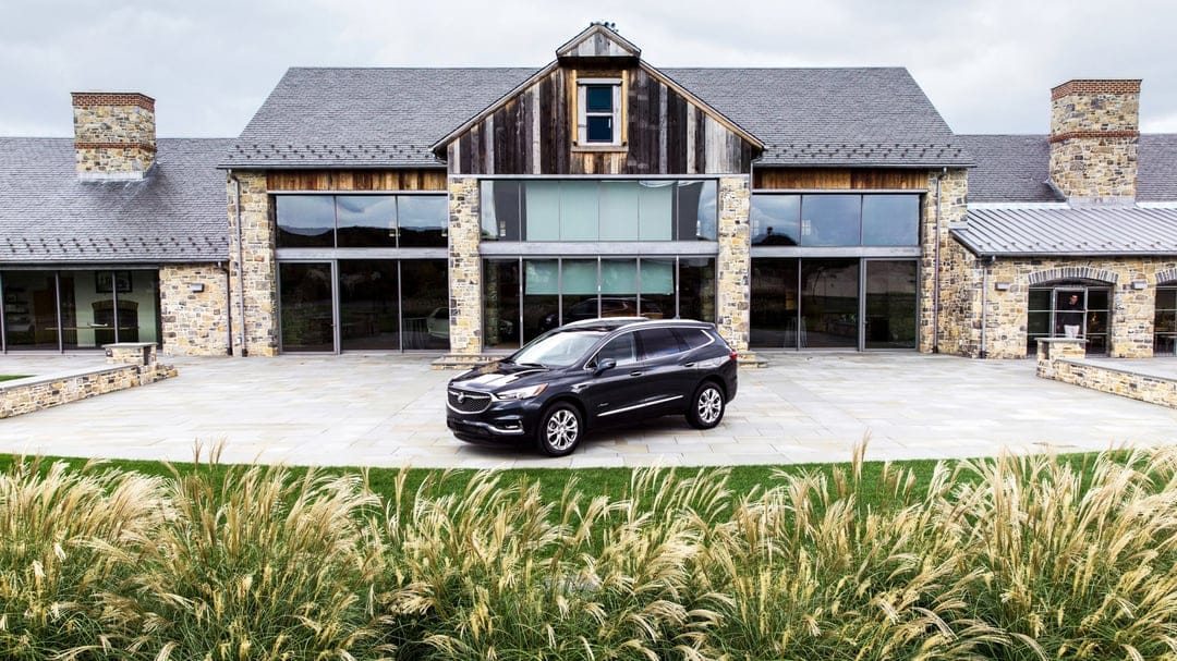 Dark grey Buick Enclave SUV parked on a stone patio in front of a modern wood and stone estate with large windows.