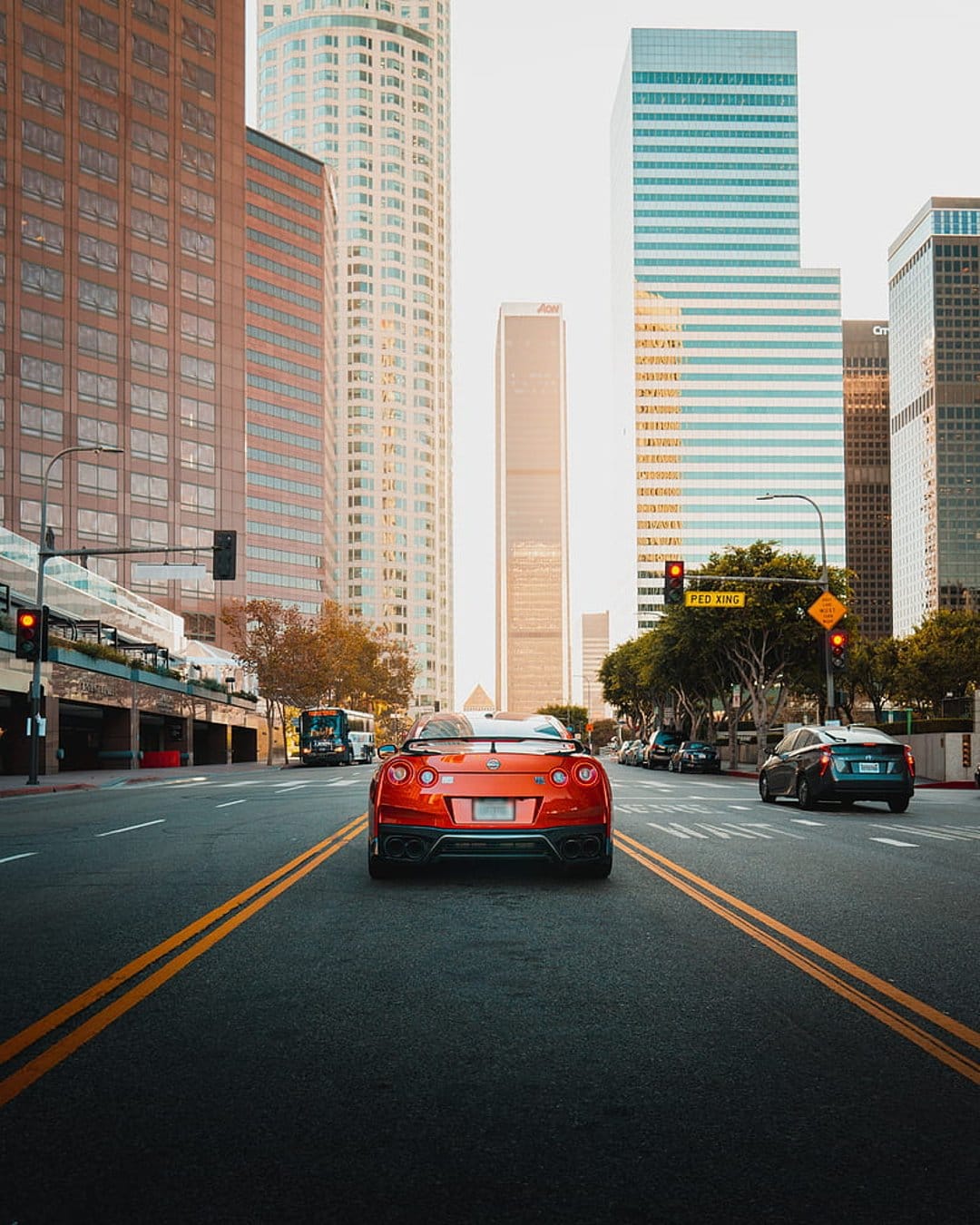 Orange sports car driving on a sun-drenched city street lined with skyscrapers and trees during golden hour.