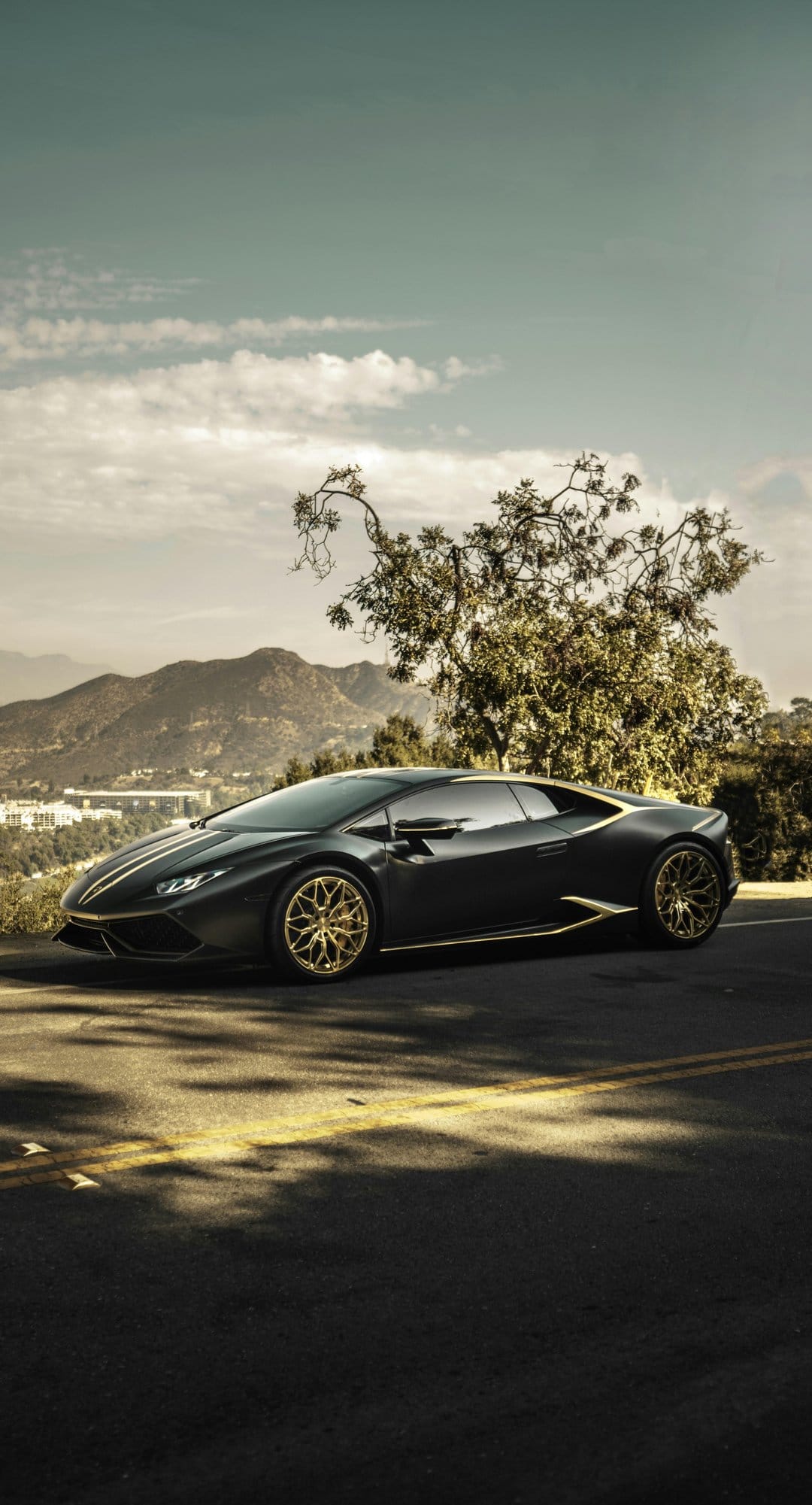Black Lamborghini with gold trim parked on a winding mountain road under a cloudy sky during the daytime.