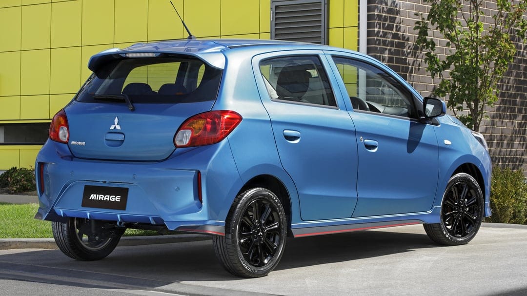 Blue Mitsubishi Mirage hatchback with black wheels parked in front of a yellow panel and brick wall.