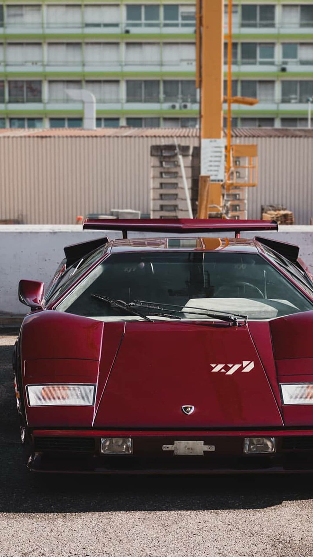 Maroon Lamborghini Countach with pop-up headlights parked on gravel near an industrial crane and tall building.
