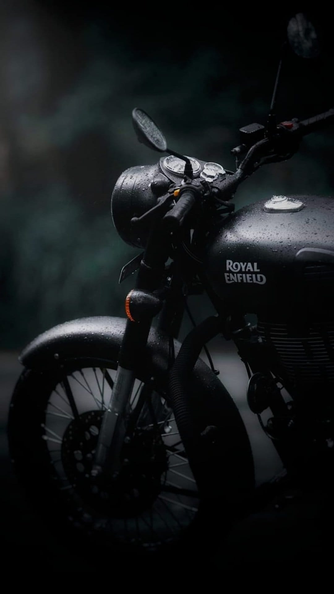 Black Royal Enfield motorcycle with water droplets on the headlight parked against a dark blurred forest.
